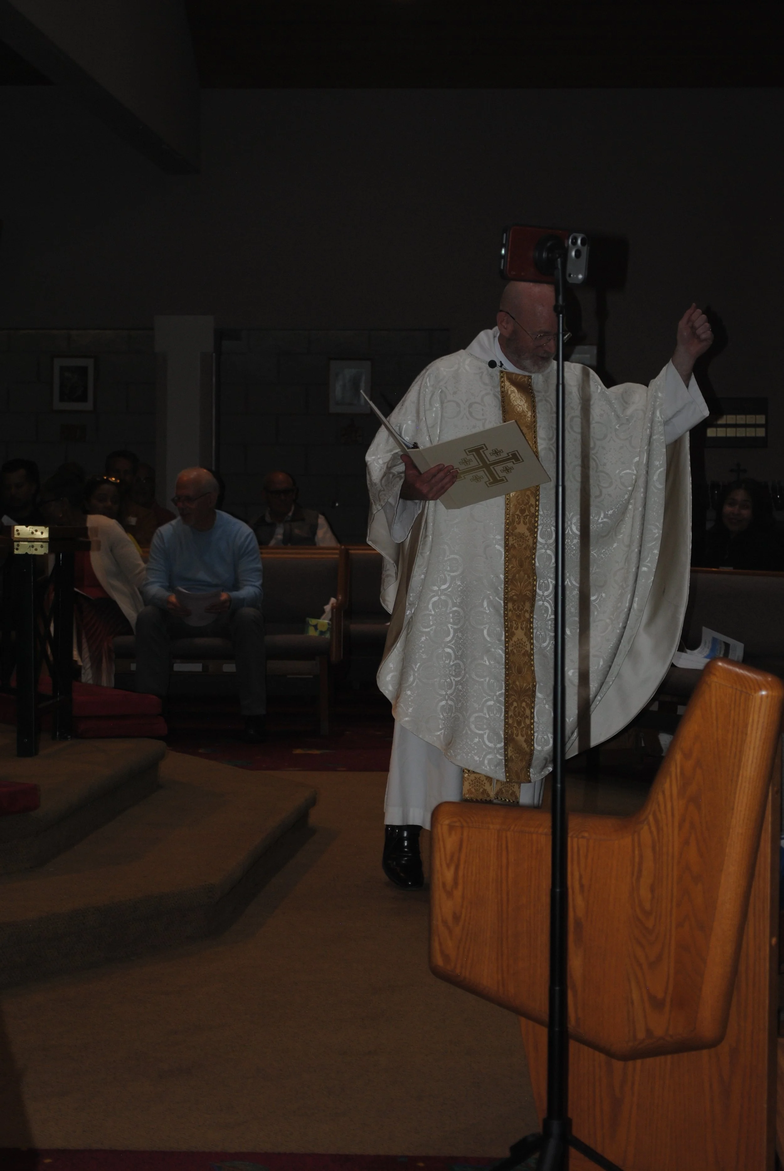 A priest in white and gold robes stands behind a wooden lectern, holding a book with a Christian cross on the cover during a church service. Several people are seated in the pews in the background.