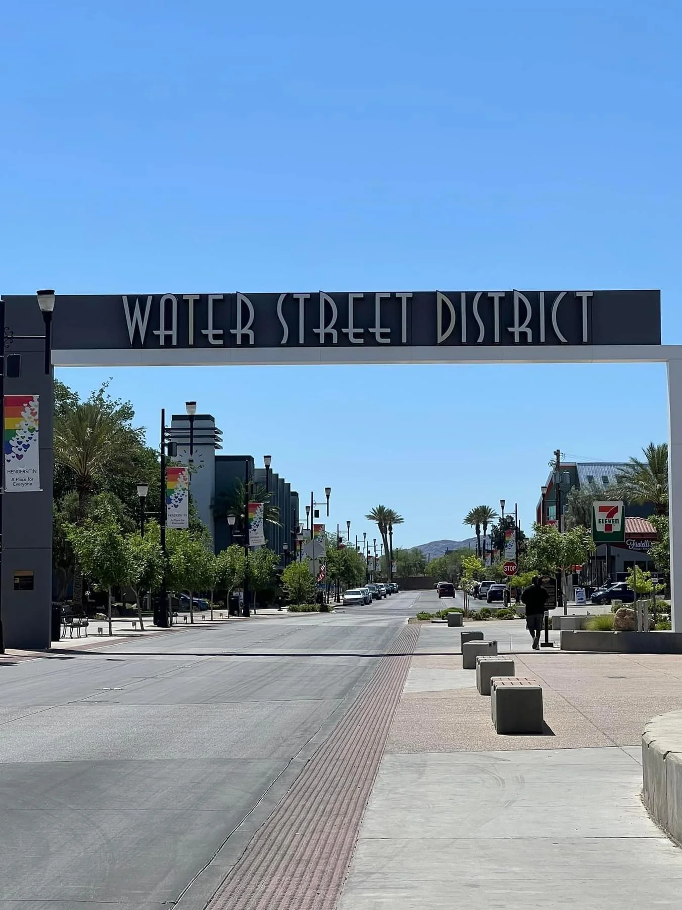 A shopping district called Water Street District with trees, lampposts, and storefronts including a 7-Eleven. The street is mostly empty with a few people and cars, under a clear blue sky and palm trees in the background.