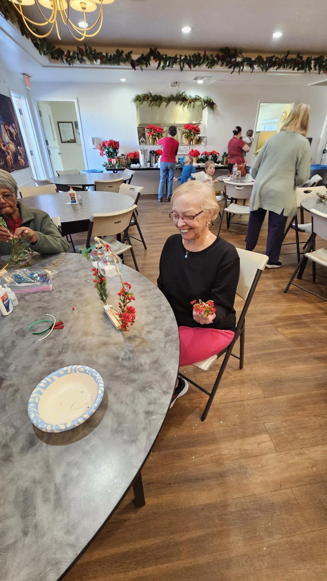 A group of people participating in a holiday craft activity in a decorated room, with Christmas-themed decorations, poinsettias, and a festive atmosphere.