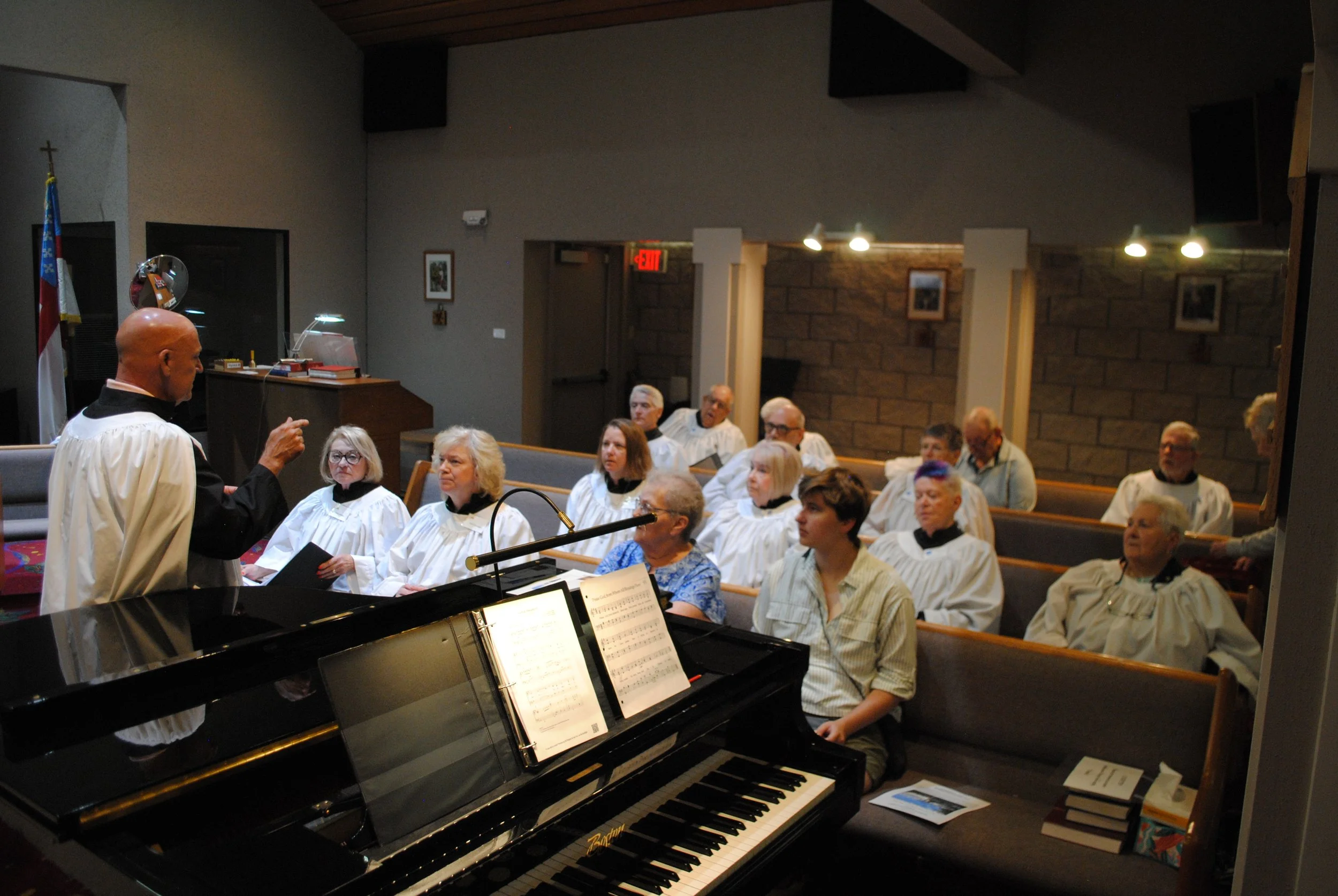 A choir of children and adults dressed in white robes sitting in pews inside a church, participating in a service or concert, with a man playing a piano in the foreground.