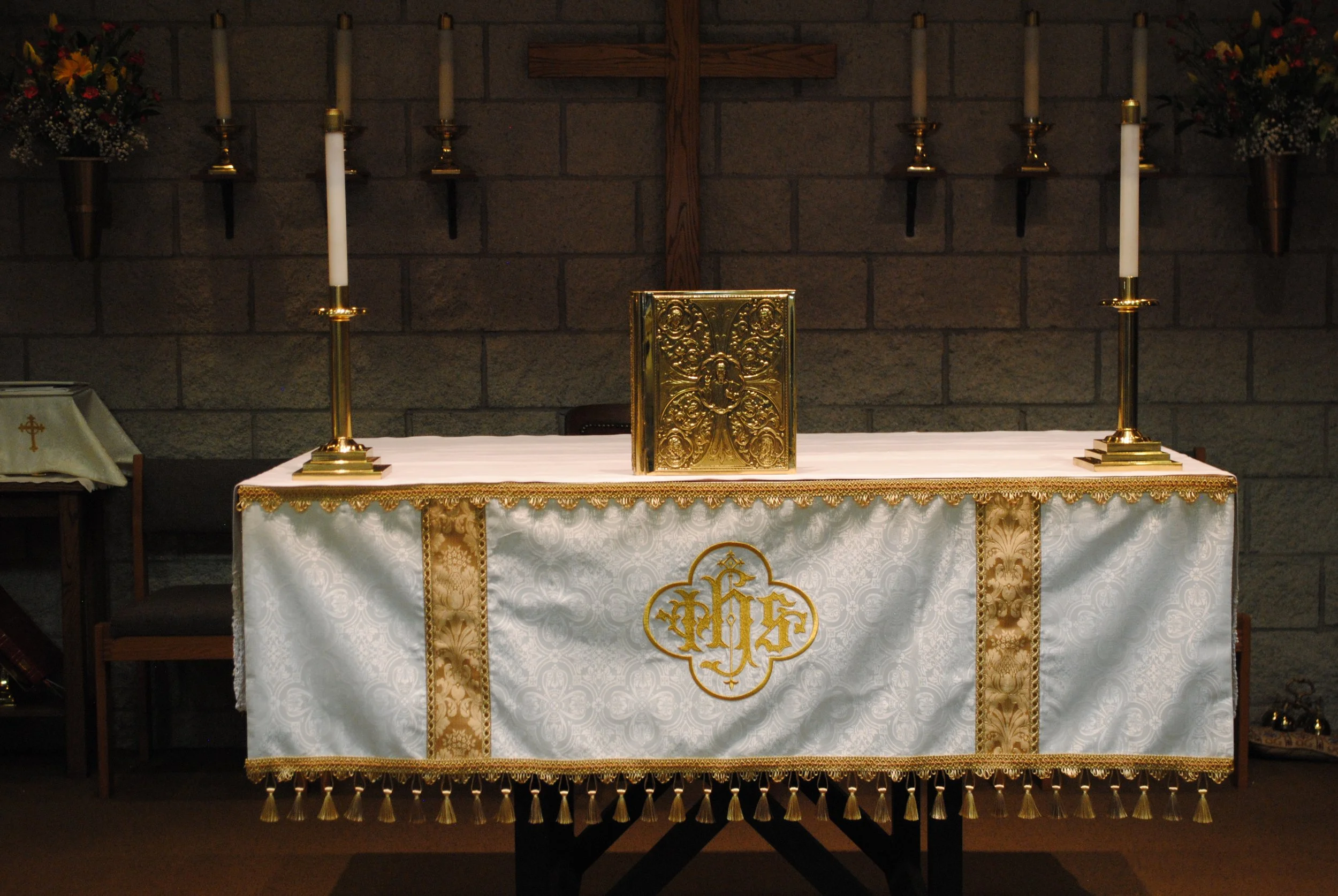 A church altar with a white cloth featuring gold embroidery, two gold candlesticks with white candles, a gold ornate Bible on a stand, and a cross on the stone wall behind. There are floral arrangements on either side.