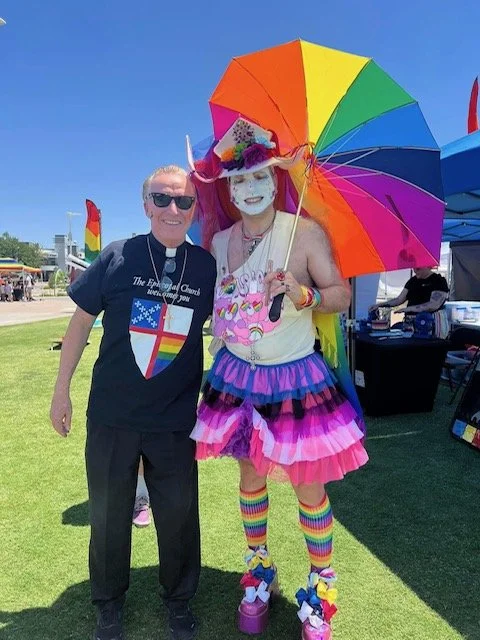 A man and a performer dressed in colorful, rainbow-themed attire at an outdoor event, with the performer holding a rainbow umbrella.