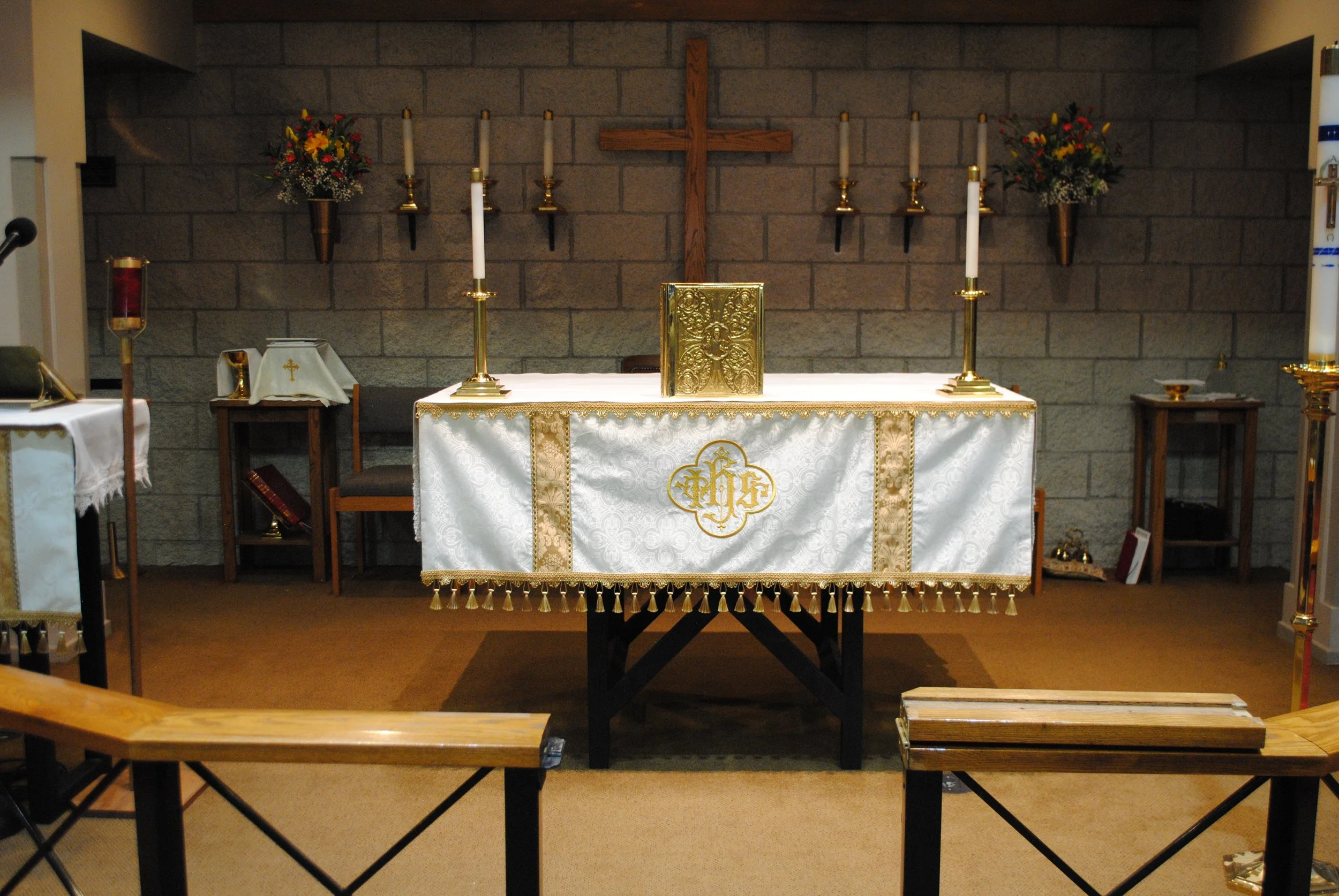 An altar inside a church with a wooden cross on a brick wall, floral arrangements, seven white candles in gold holders, a gold tabernacle, and a white altar cloth with gold embroidery.