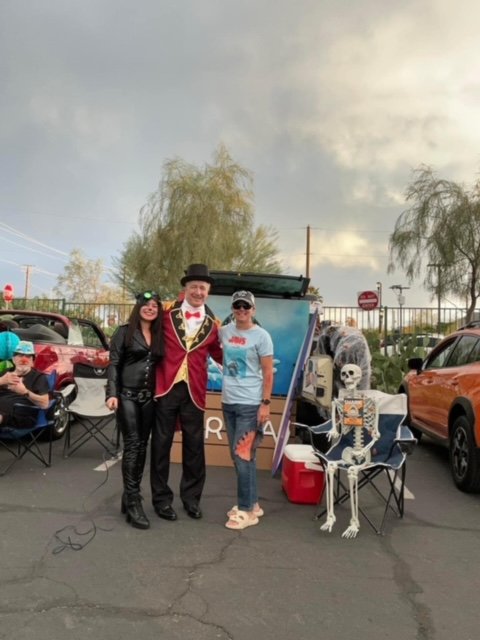 Three people standing outdoors in a parking lot, dressed casually and semi-formally, with Halloween decorations including a skeleton sitting on a chair, a large skull, and a painted backdrop. There are parked cars and trees in the background.