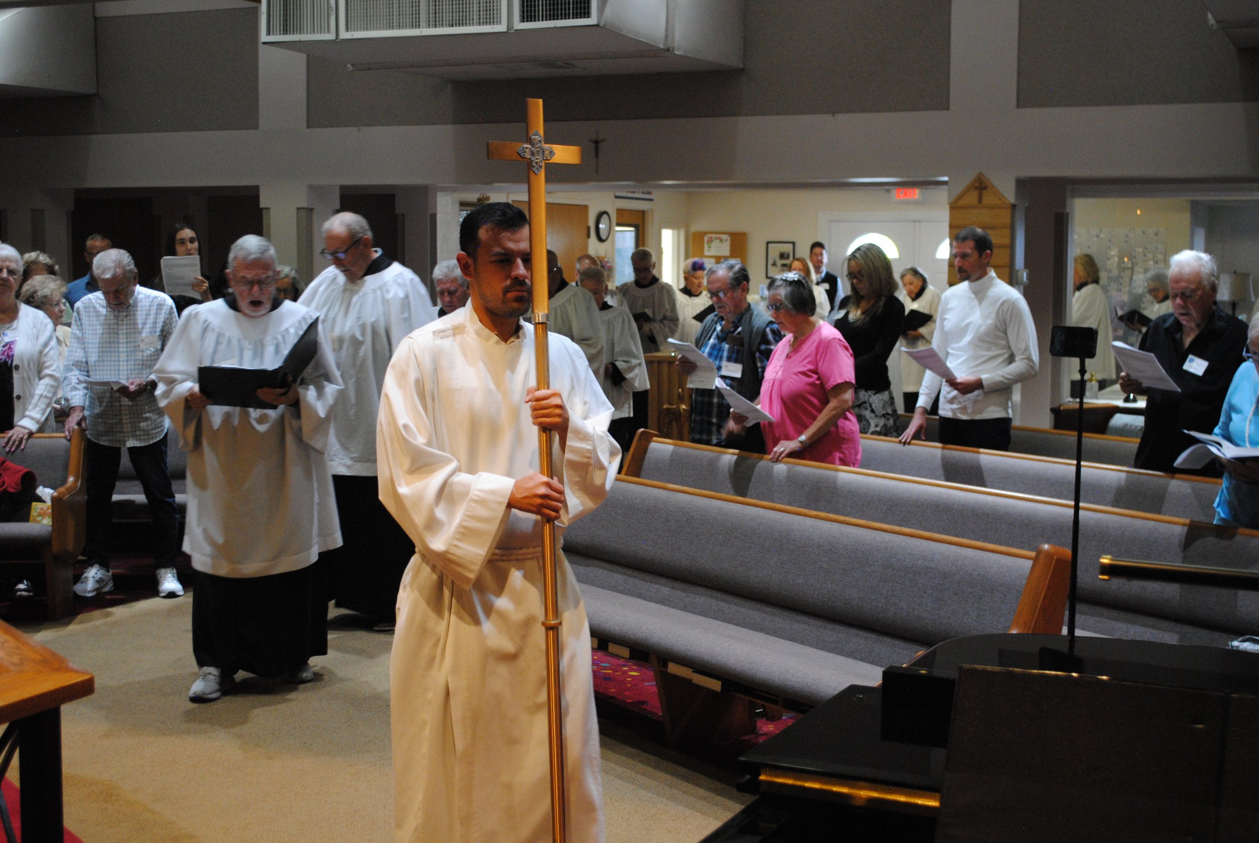 A religious ceremony in a church showing a person in a white robe holding a cross, surrounded by congregants, some of whom are singing or reading from booklets.