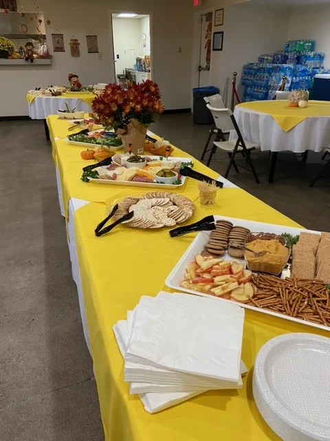 Buffet table with snacks and appetizers, including sandwiches, cookies, and crackers, decorated with a flower arrangement, in a room with water bottles and additional tables in the background.