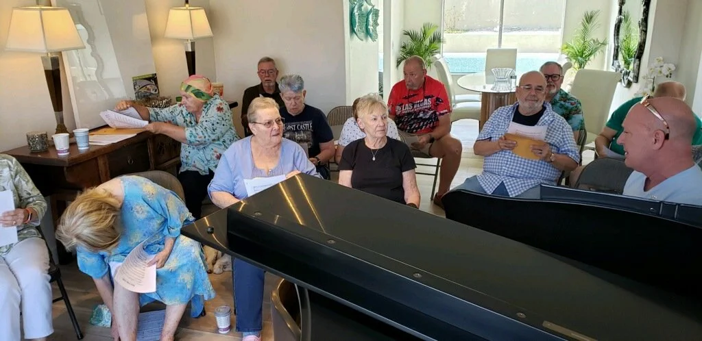 Group of elderly and middle-aged people gathered around a black piano, singing or participating in an activity in a bright, airy room with large windows and plants.