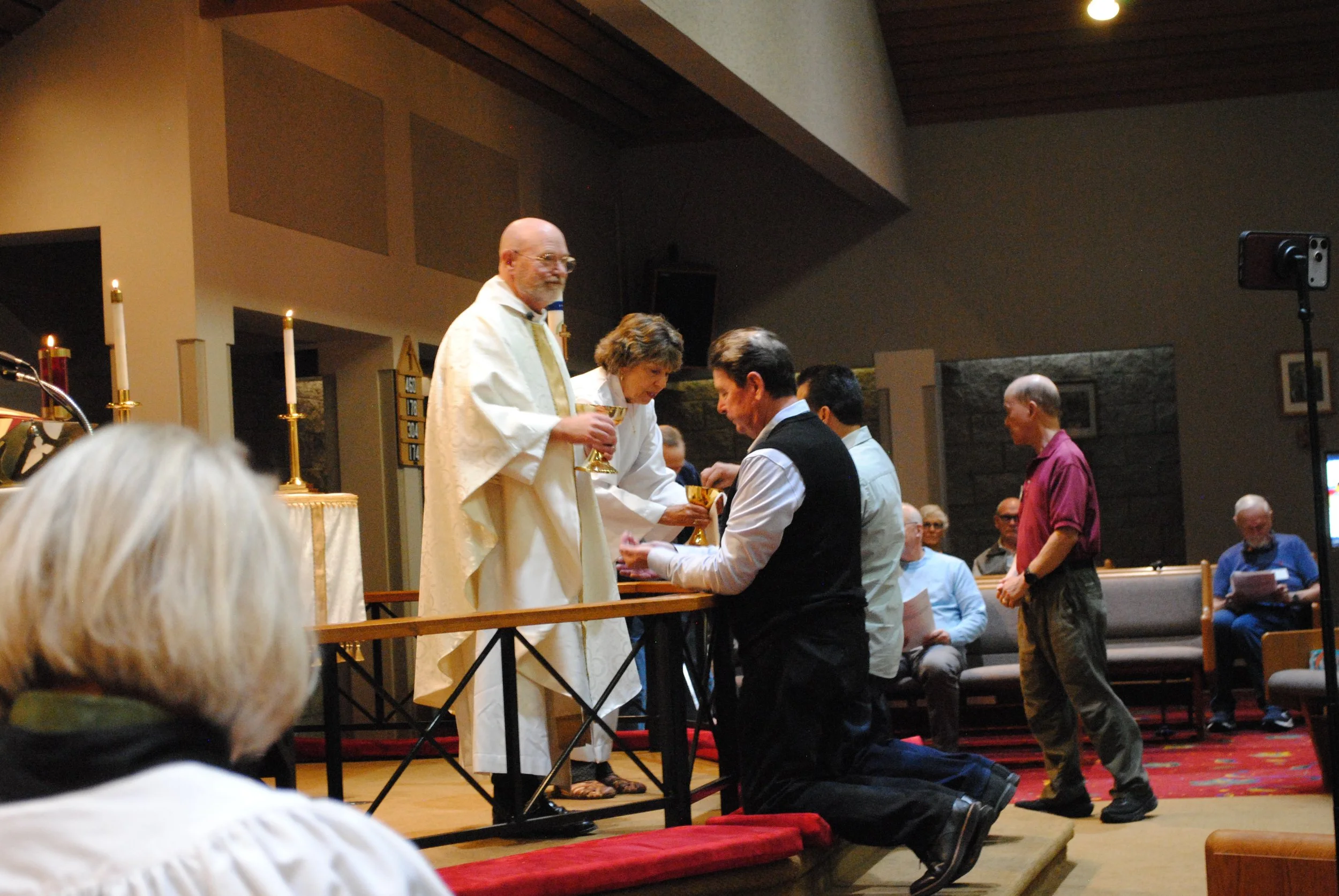 A church service or religious ceremony taking place inside a church, with priests or clergy administering communion or blessings to congregants. People are kneeling and receiving communion at the altar, which has candles and religious items.