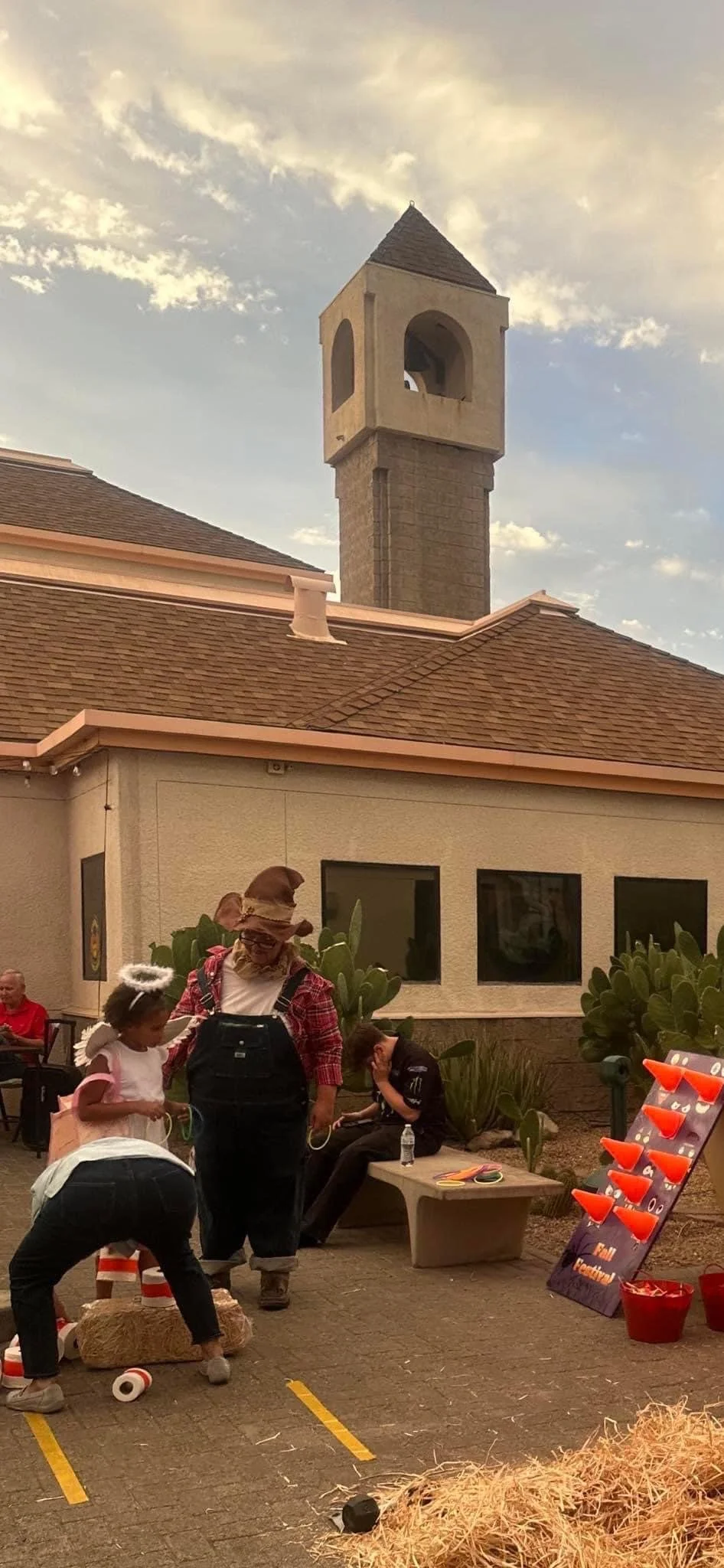 People gathered outside a building with a clock tower, dressed in costumes, including an angel and a scarecrow, with Halloween-themed decorations.
