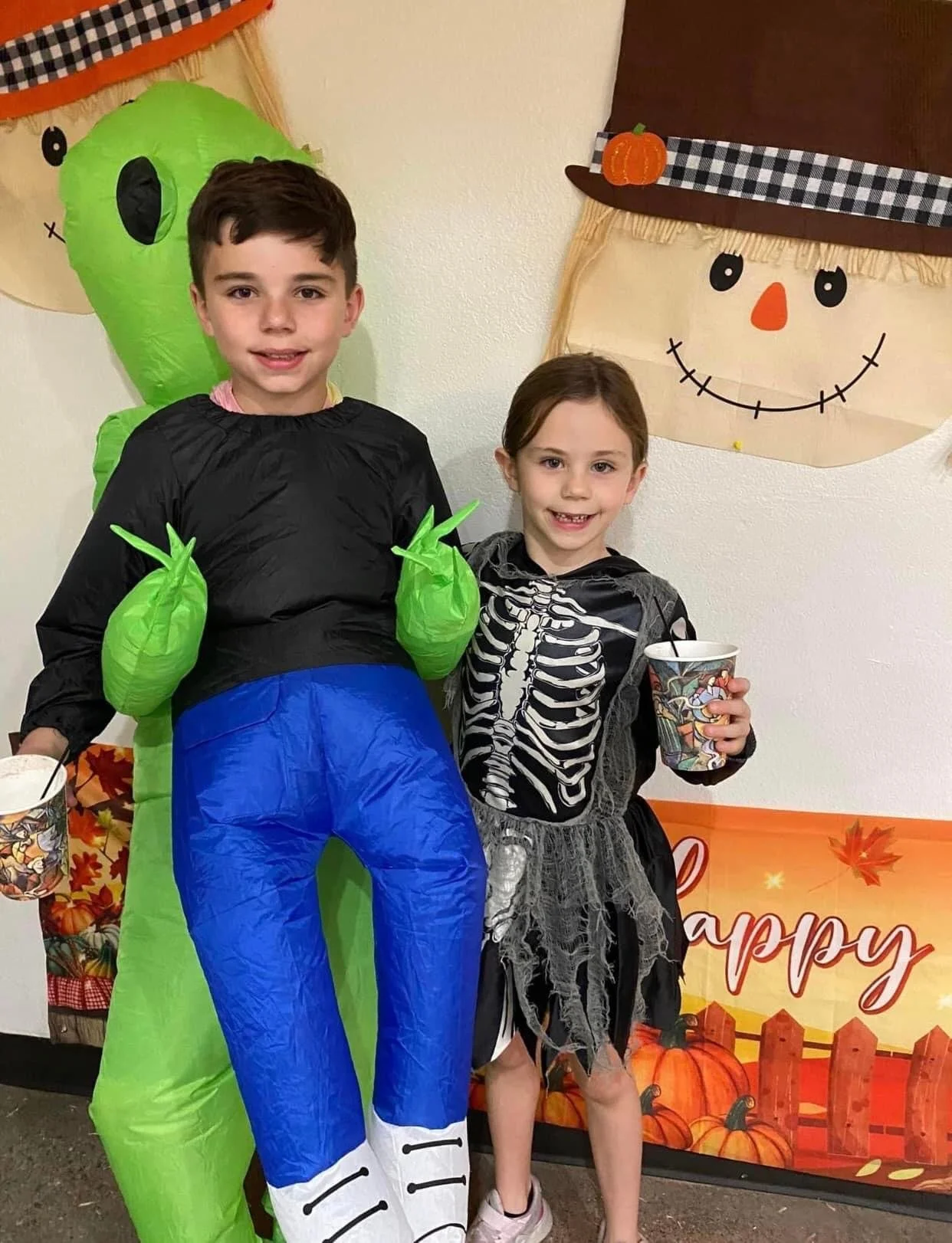 Two children dressed in Halloween costumes standing in front of fall-themed decorations. The boy is dressed as a green alien with green gloves and an inflatable body suit. The girl is dressed as a skeleton with a black dress and a skeleton print. She