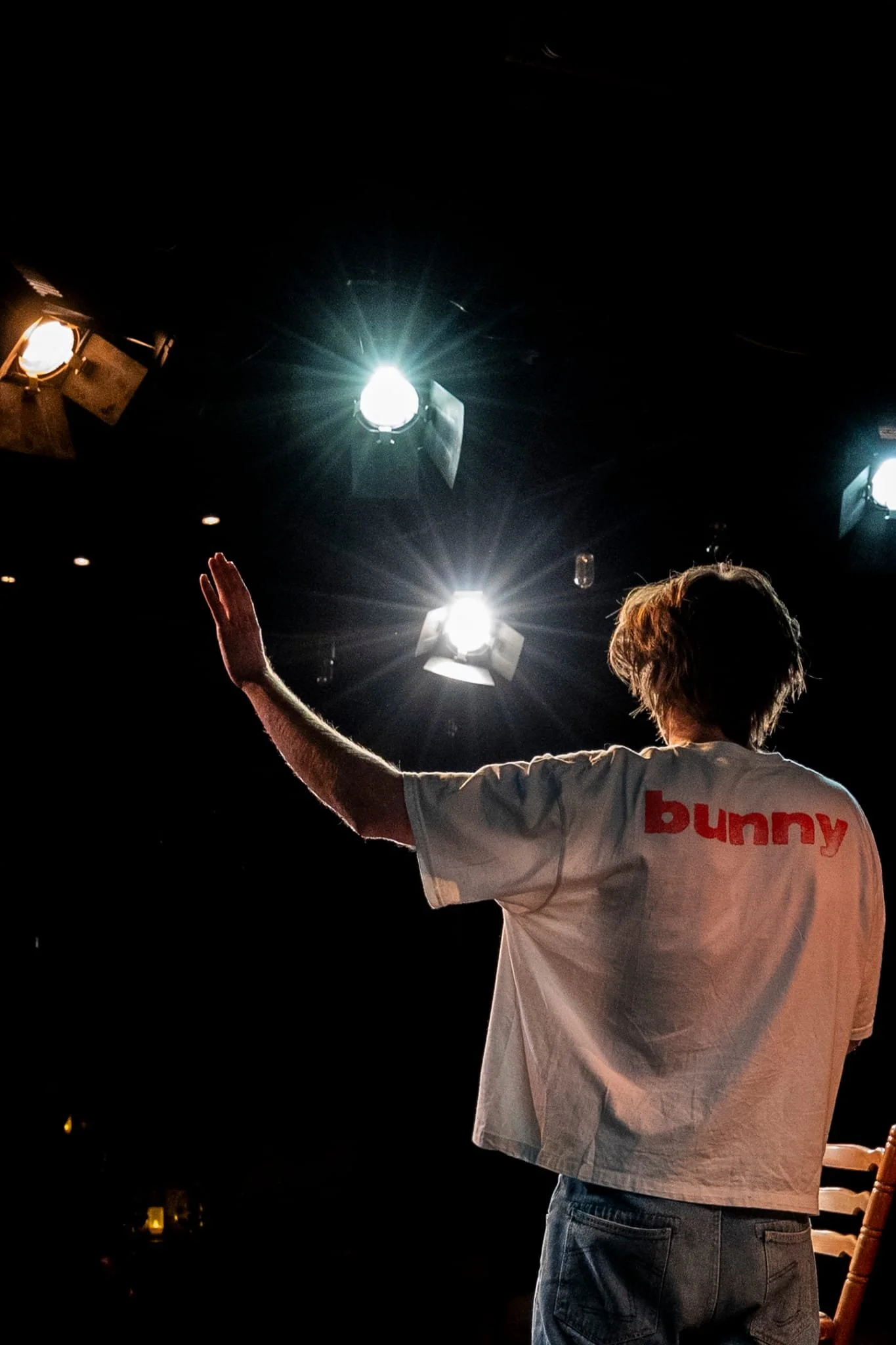 Een persoon met bruin haar, gekleed in een witte T-shirt met de tekst 'bunny' op de rug, zwaait met één hand naar de camera. De achtergrond is donker met heldere studiolampen die op de persoon gericht zijn.