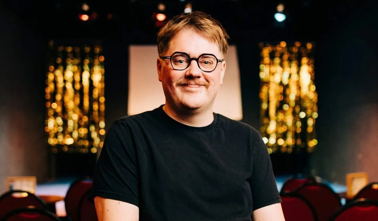 Bart Michiels, voorzitter  en mede oprichter van Nonkel Gaston. Man met bril, korte haren, in een zwarte T-shirt, zittend in een theater of cabaret setting met rode stoelen en gouden decoraties op de achtergrond.