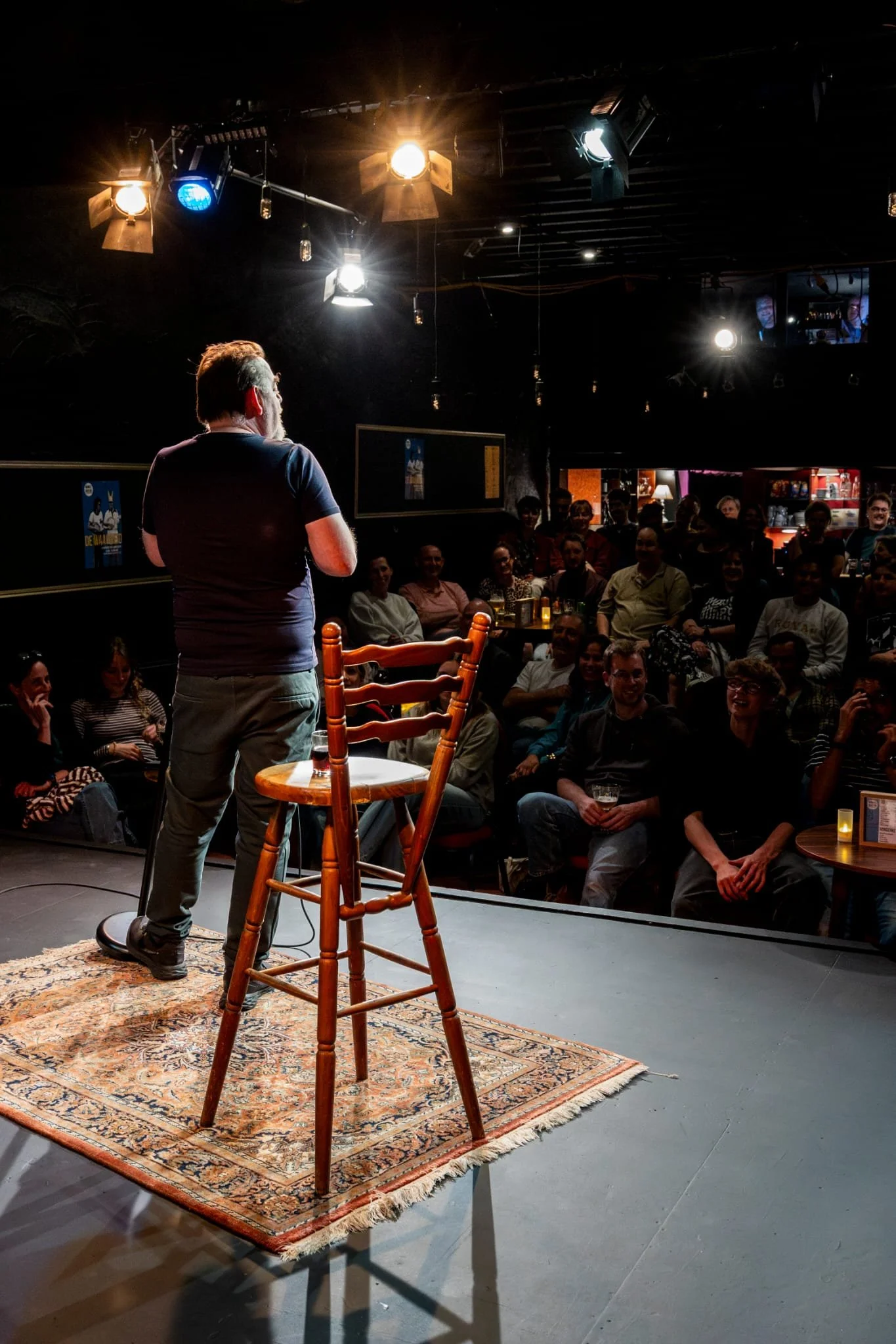 Man vertelt op een podium in een theater of comedy club, met een publiek dat lacht en plezier heeft, onder heldere verlichting.