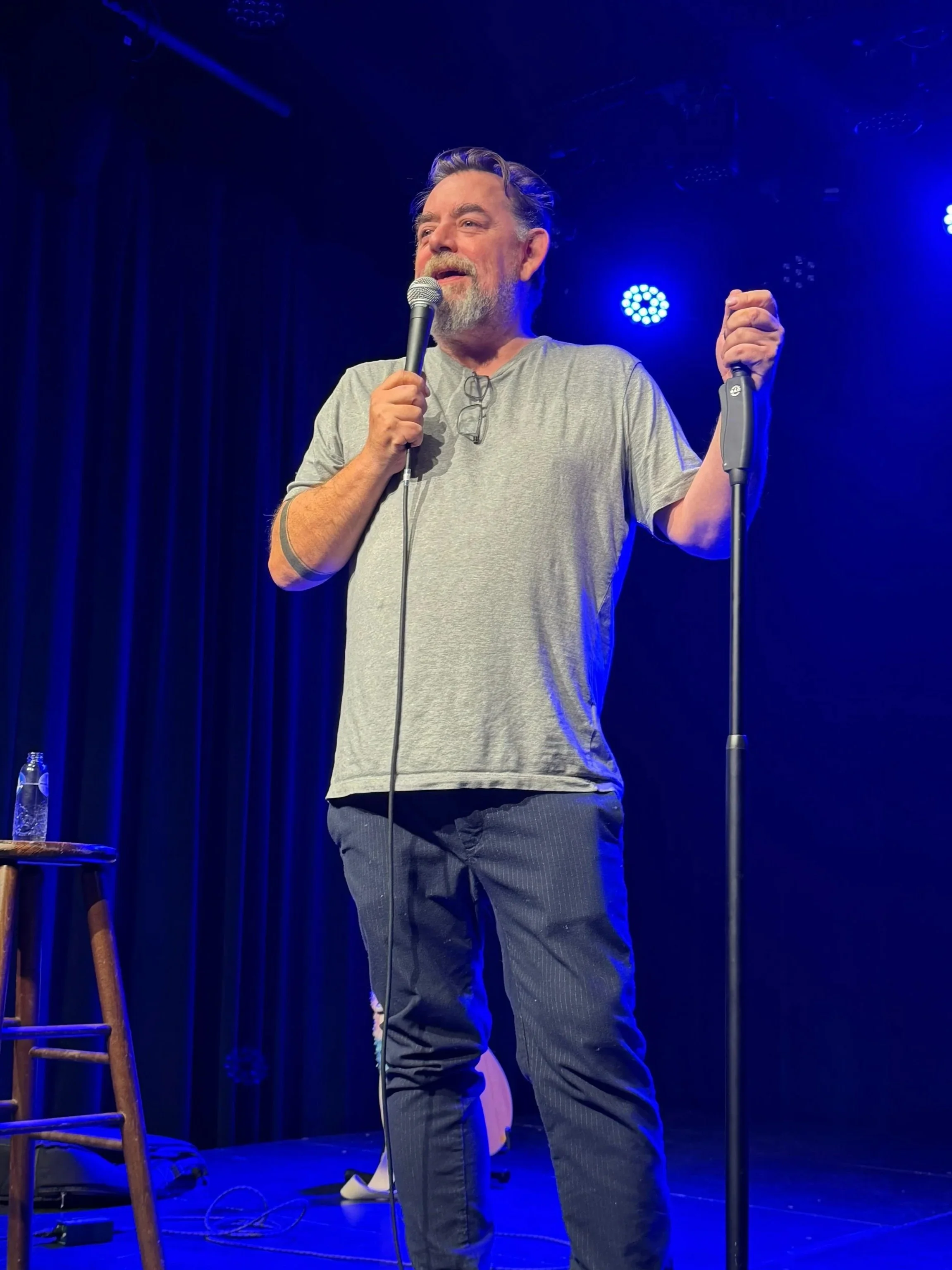 Man op het podium, houdt een microfoon in de rechterhand, blauwe achtergrondverlichting, optreedt mogelijk tijdens een comedyshow of voorstelling.