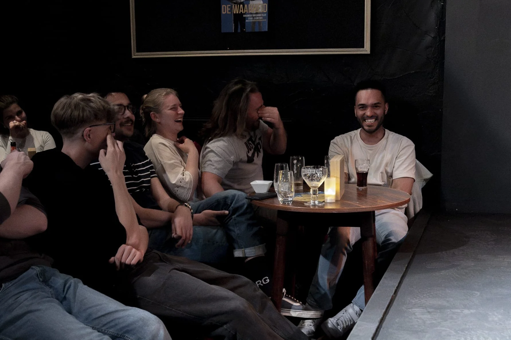 Mensen die lachen en plezier hebben tijdens een avond in een donker restaurant of café, zittend aan een ronde tafel met drankjes en kaarslicht.