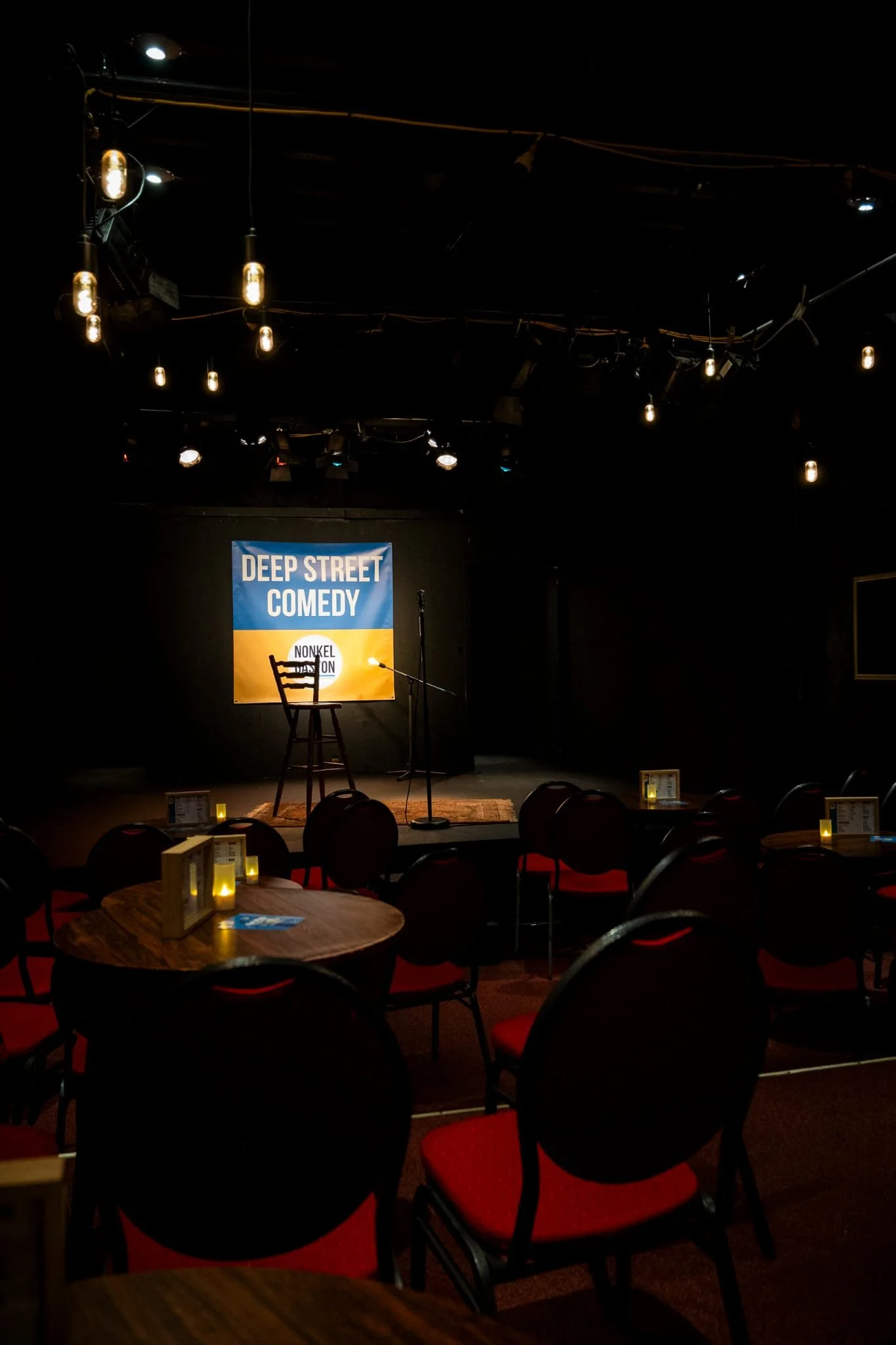 Podium met een stoel en microfoon, banner met tekst 'Deep Street Comedy' op een donkere comedyclub. Tafels met kaarsen en menukaarten in de voorgrond.