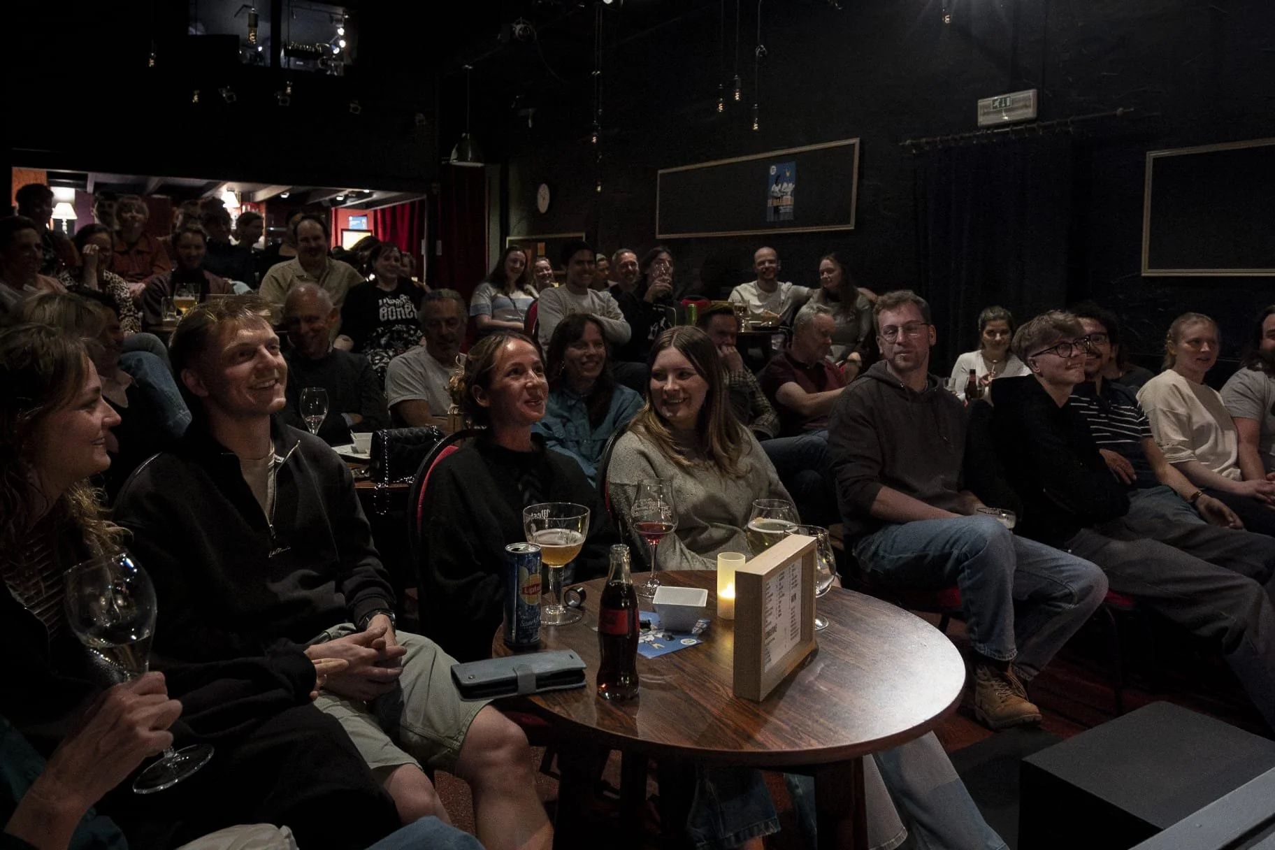 Een groep mensen zit in een donkerverlichte zaal, zichtbaar genietend en lachend tijdens een evenement of comedyshow, met drankjes en een menu op de tafel.