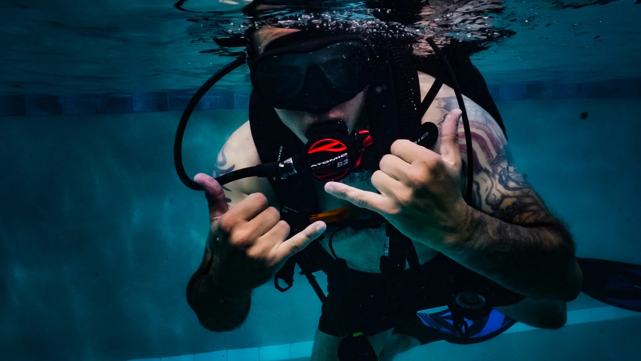 A person with tattoos on their arm underwater wearing a scuba mask and holding a regulator, giving a thumbs-up.