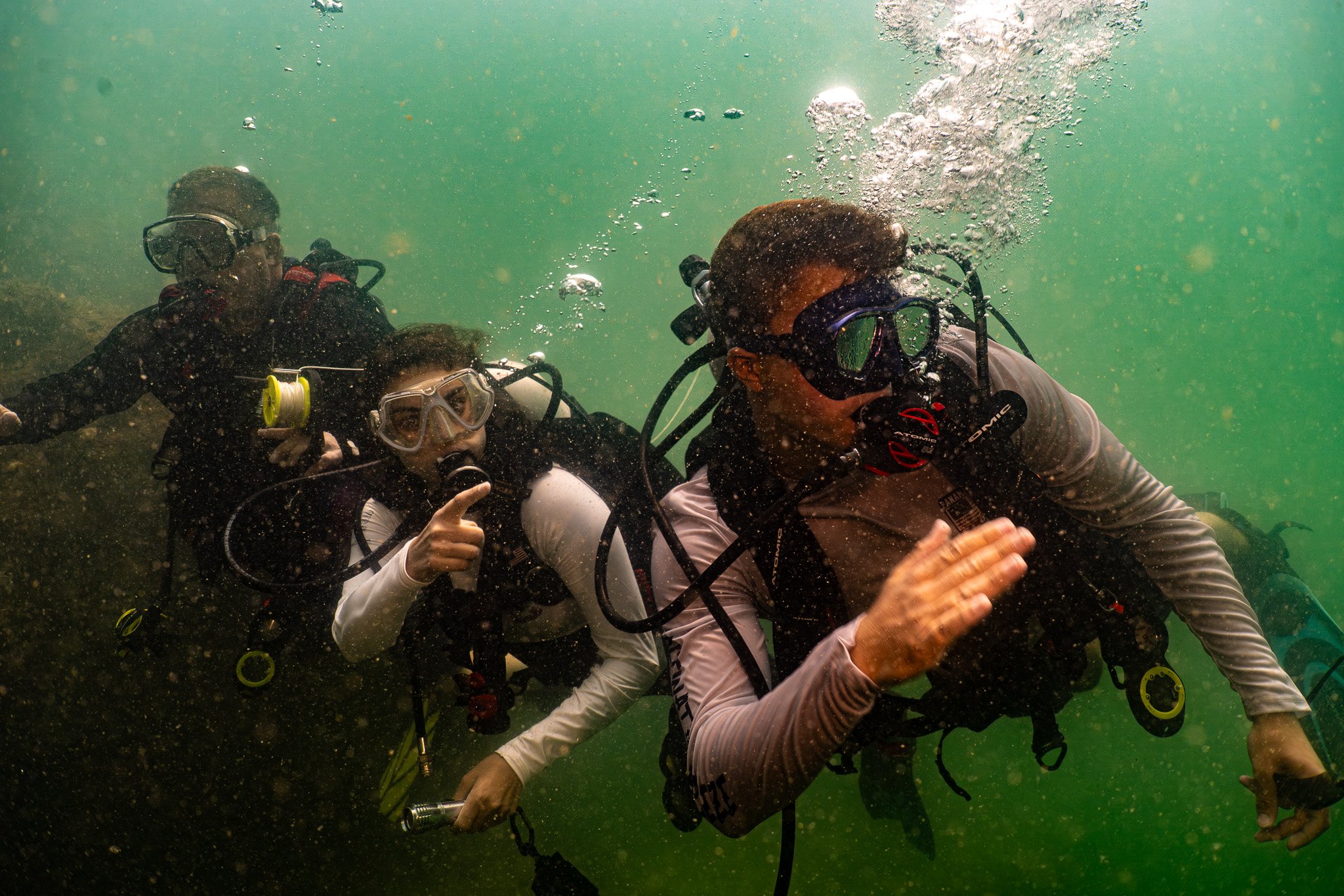 Three scuba divers underwater, wearing masks and suits, giving thumbs-up signs, surrounded by bubbles and green water.