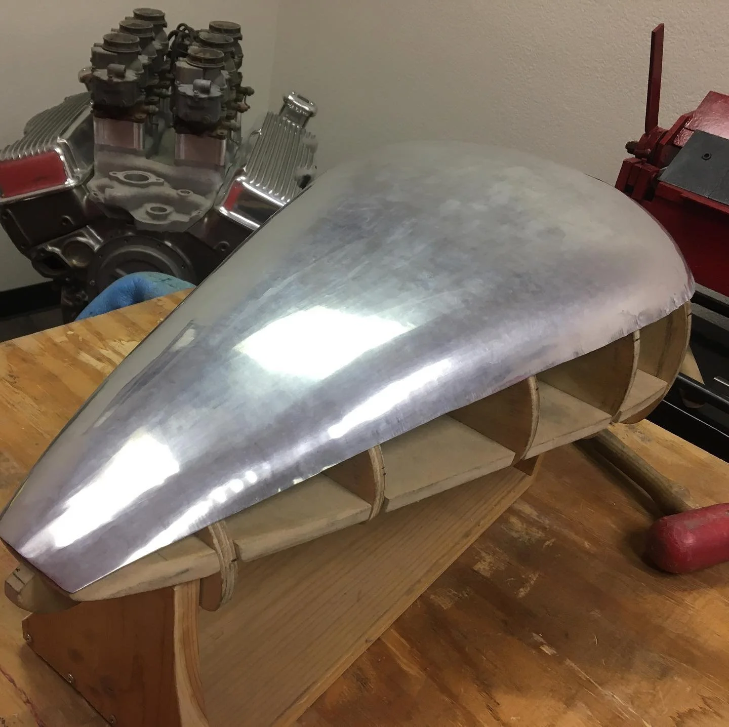 A partially assembled elongated metal airplane fuselage on a wooden workbench with a wooden frame inside. In the background, there is an engine and other tools.