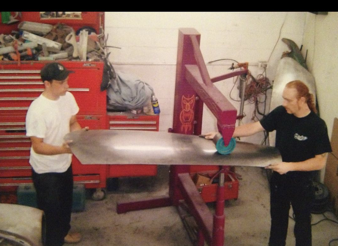 Two people handling a large, curved piece of metal in a workshop, with tools and a red tool chest in the background.