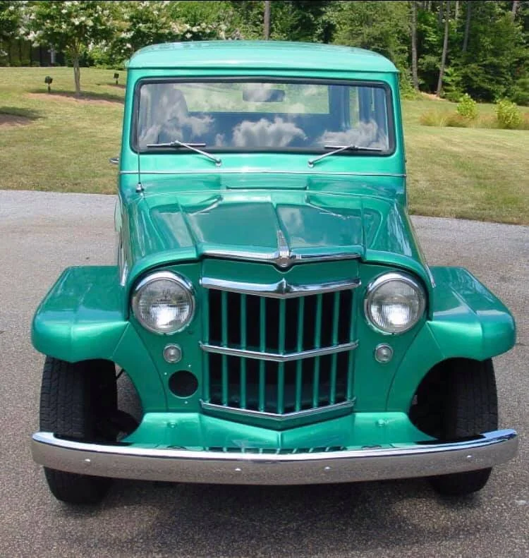 Front view of a vintage green automobile with a distinctive grille and round headlights, parked on a gravel surface with greenery in the background.