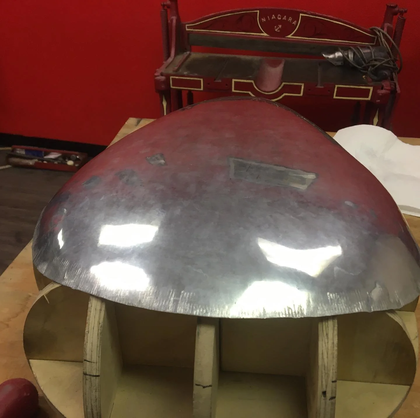 A metallic dome-shaped object with a reflective surface placed on a wooden structure indoors. In the background, there is a red Niagara-branded cart with some wires and an object on top, against a red wall.