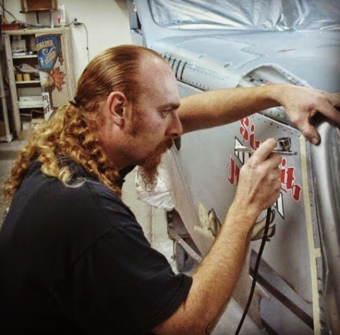 A man with long red hair and a beard is painting a sign on a vending machine in a workshop.