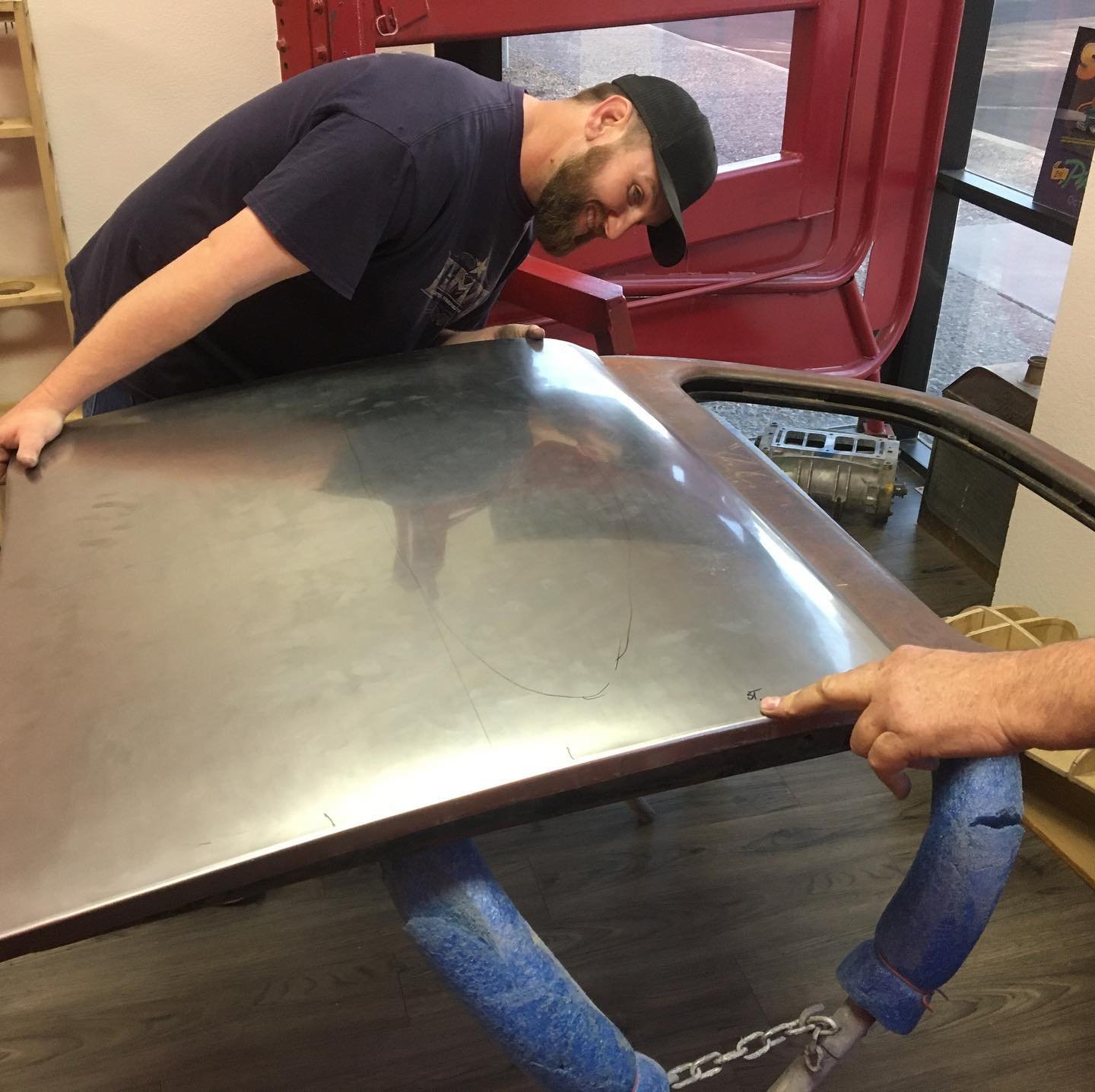 A man wearing a black cap and a dark T-shirt is looking underneath a vintage car hood, which is supported by a manicurist hand with a chain. The scene appears to be inside a store or workshop with a red folded table and a window in the background.