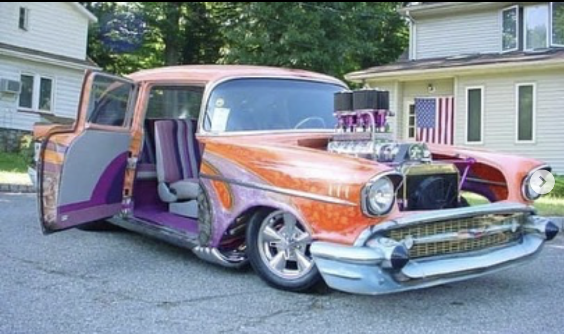 A customized vintage car with a pink, purple, and orange paint job, modified with a body kit and large engine exposed at the front, parked outside in a residential area with houses and trees in the background.