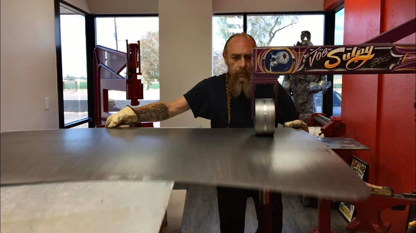 A man with a long beard and tattooed arms cutting metal with a power saw in a workshop.