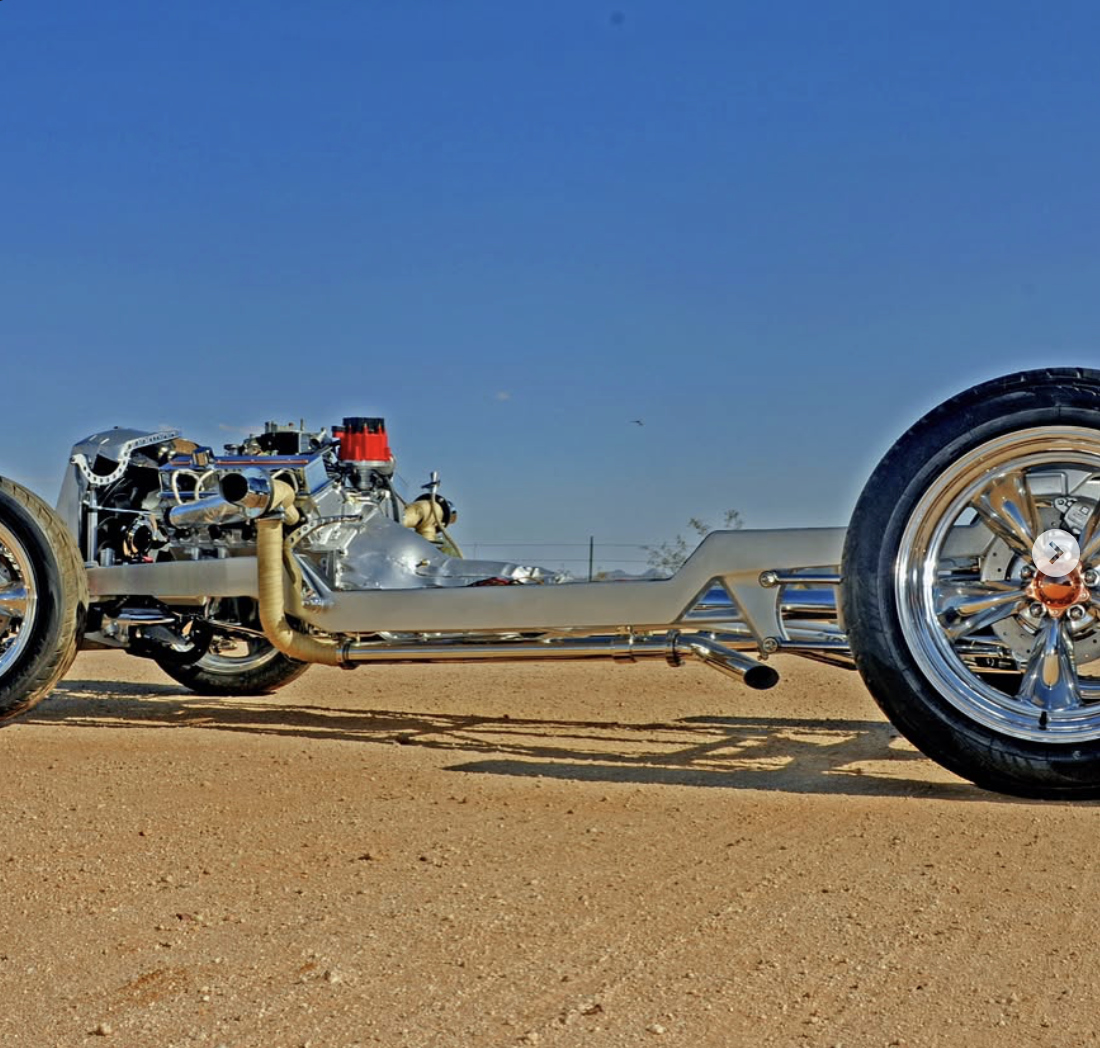 A vintage drag racing car with an exposed engine, large rear tires, and a sleek body, parked on a dirt track under a clear blue sky.