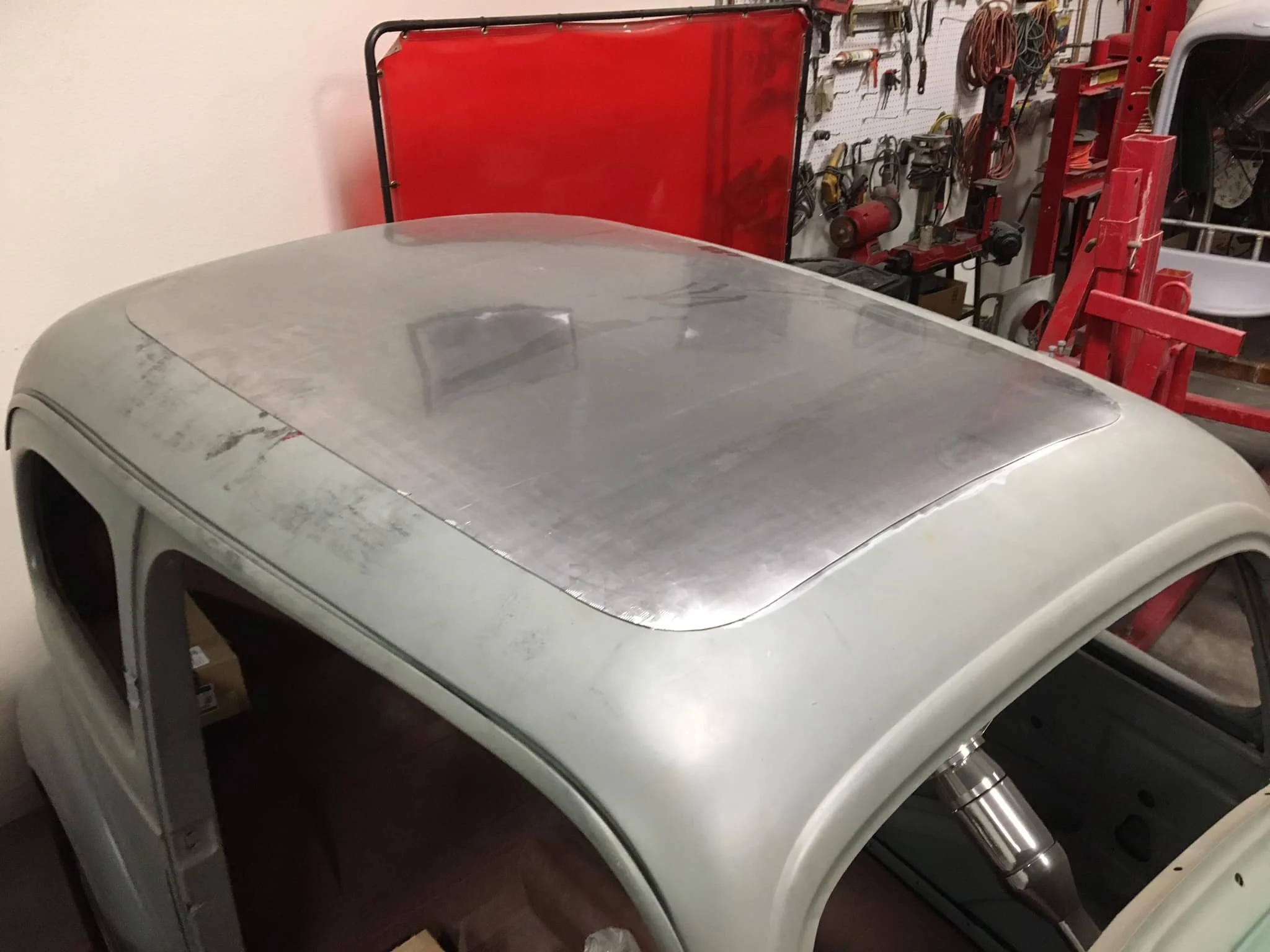 The top of a vintage car with a metal roof panel in a garage, surrounded by tools and equipment hanging on a pegboard and stored on shelves.