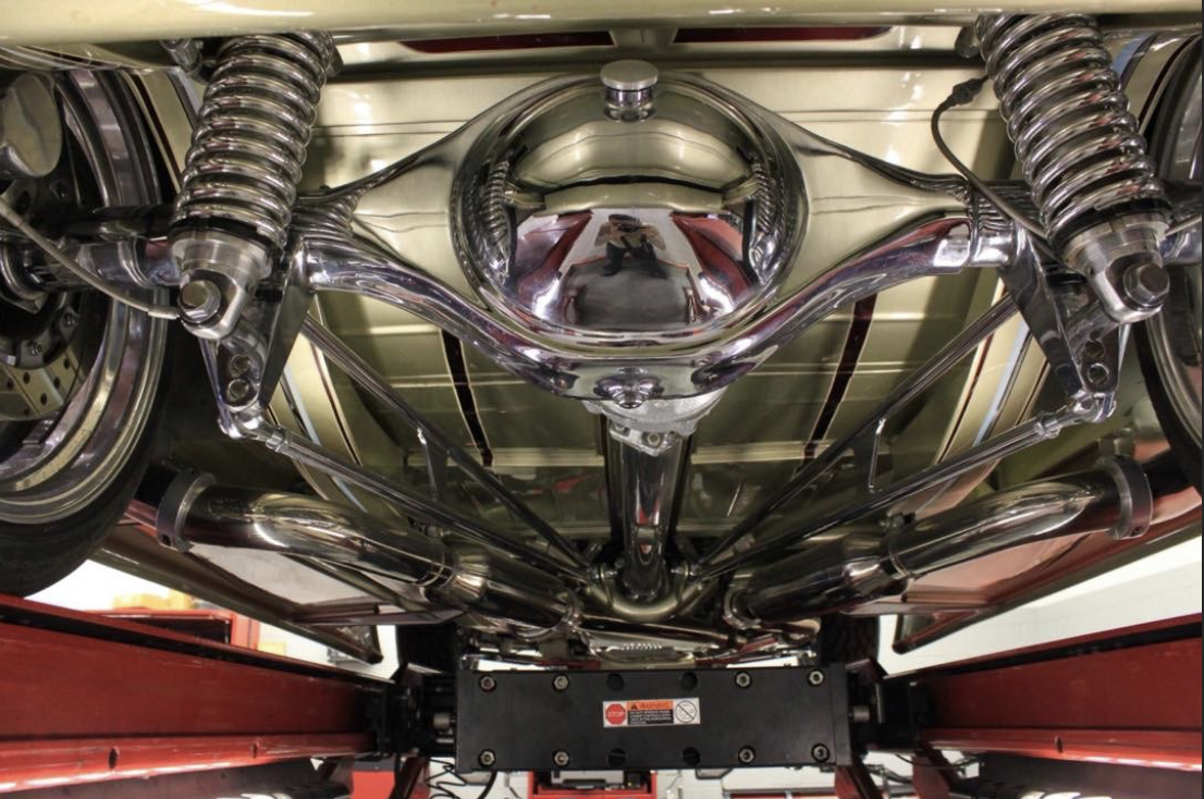 The underside of a vintage motorcycle showing shiny metal components, exhaust pipes, suspension, and wheels.