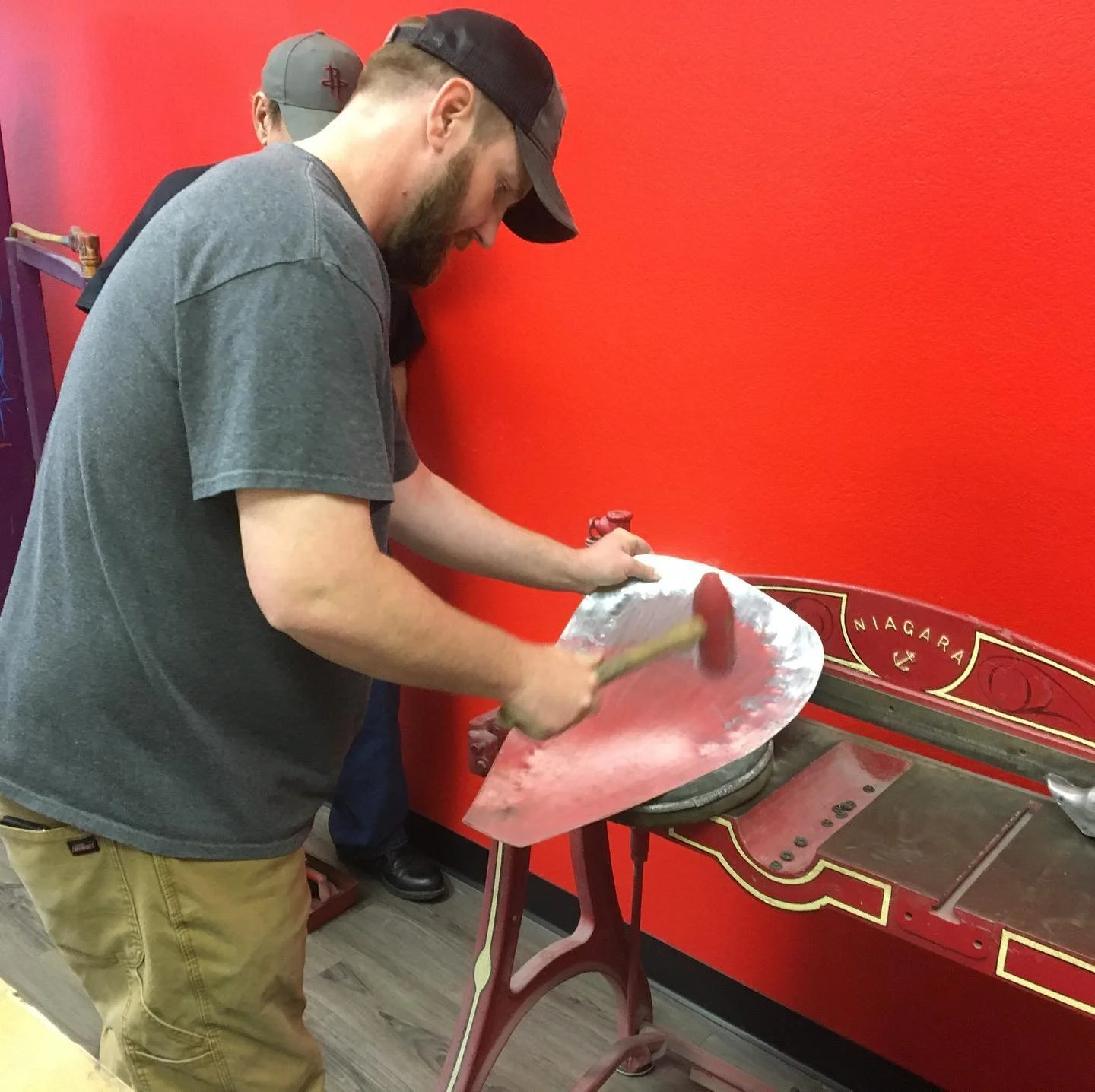 A man pounding a metal hockey puck on a vintage Niagara-brand anvil, with a red background, inside a room.