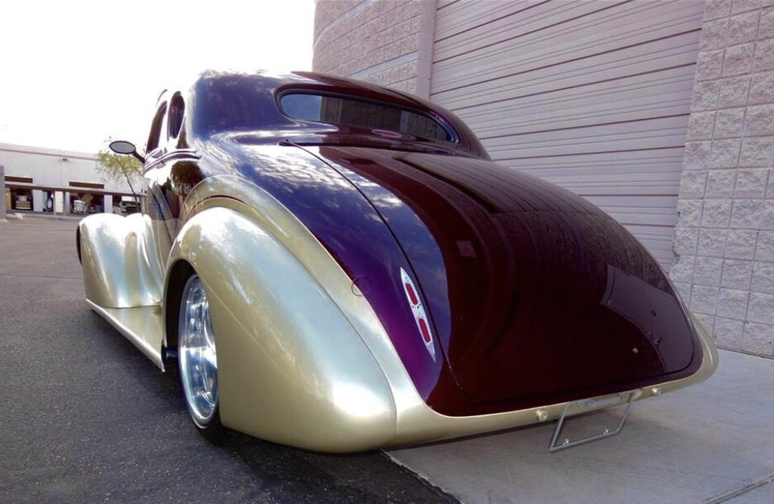 A vintage custom car with a two-tone paint job, part black and part cream, parked in front of a building with a sidewalk and large garage door.