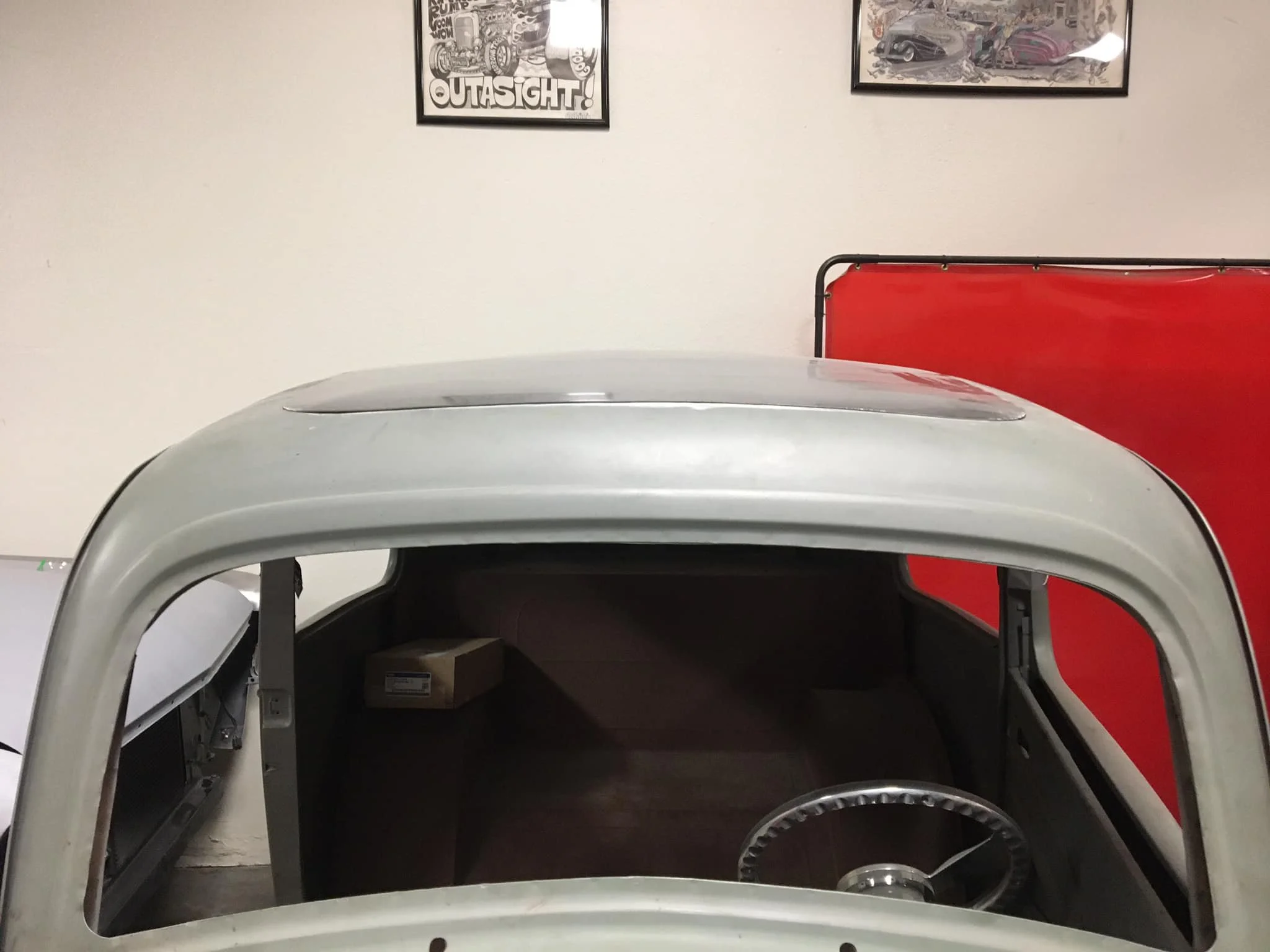 View from above of the interior of a vintage car shown from the windshield with the steering wheel visible, with framed posters of cars on the wall behind it.