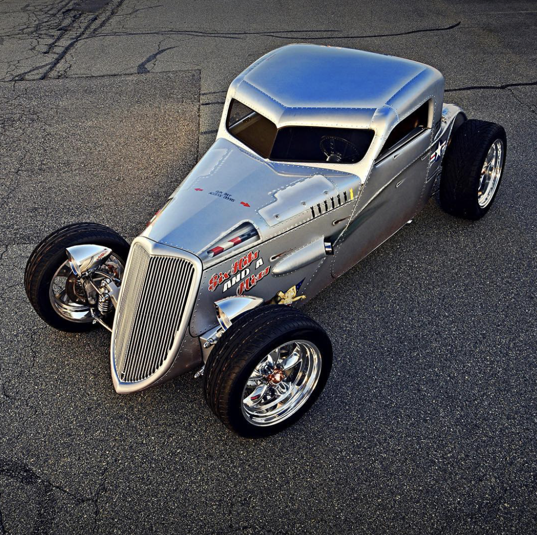 A vintage, highly polished silver racing car with exposed engine parts, wide tires, and racing decals, parked on a paved surface.