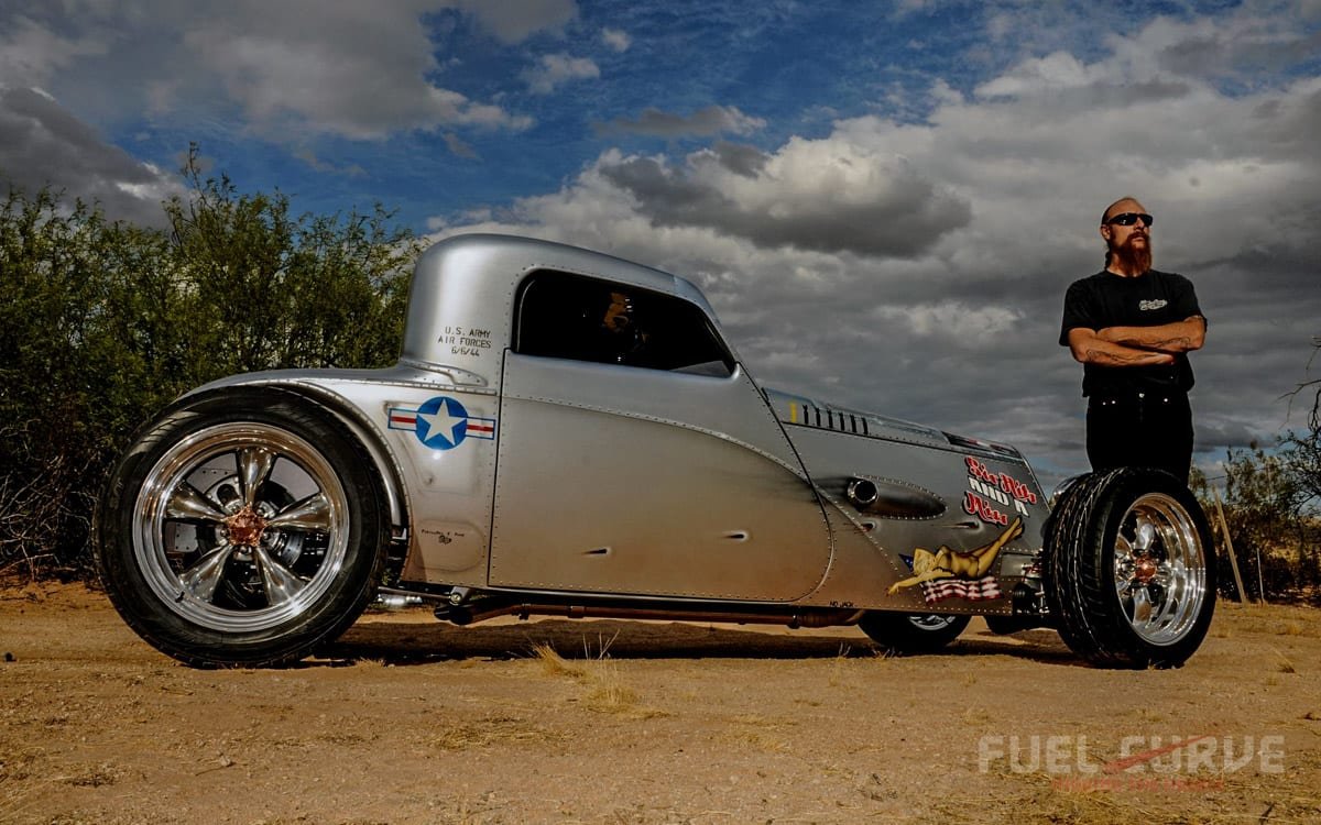A vintage-style, silver drag racing car with large rear tires and custom wheels, parked on dirt with a man in black shirt, sunglasses, and a beard standing nearby with arms crossed. The car has military markings and artwork, and the sky is partly clo
