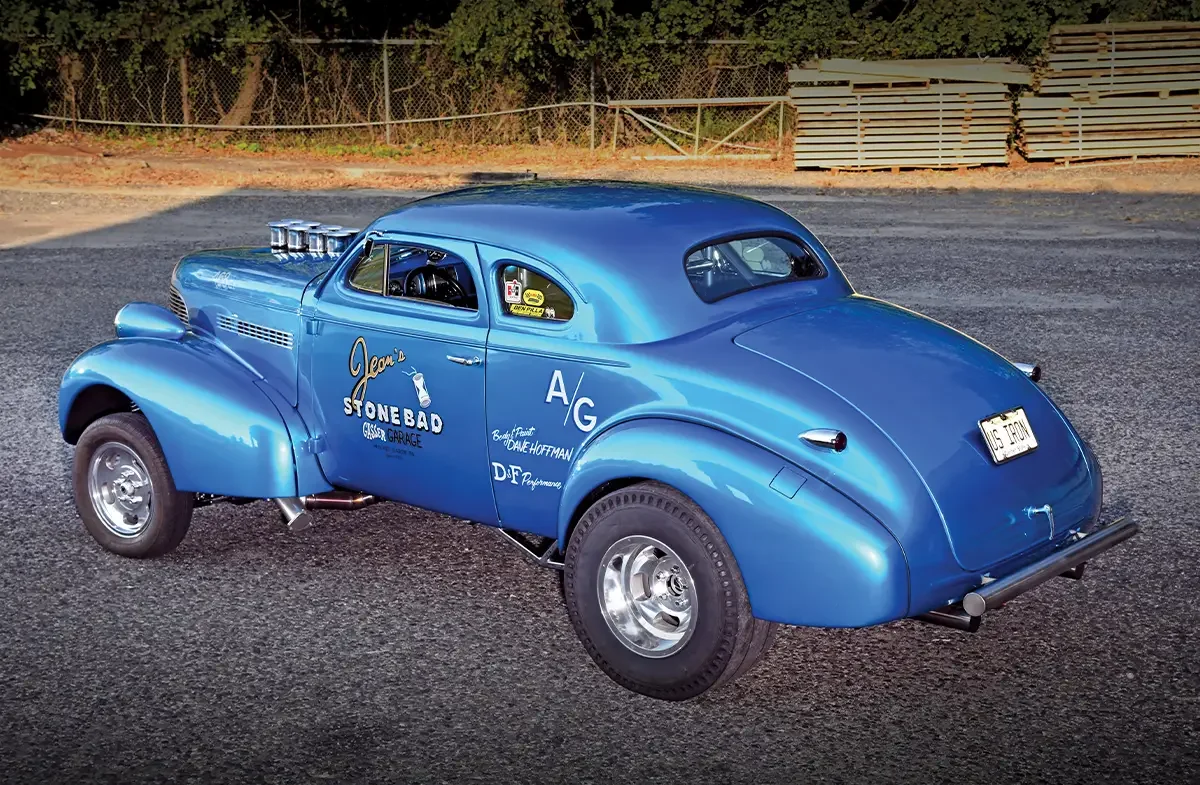 Vintage blue racing car parked on a gravel lot with trees and wooden pallets in the background.