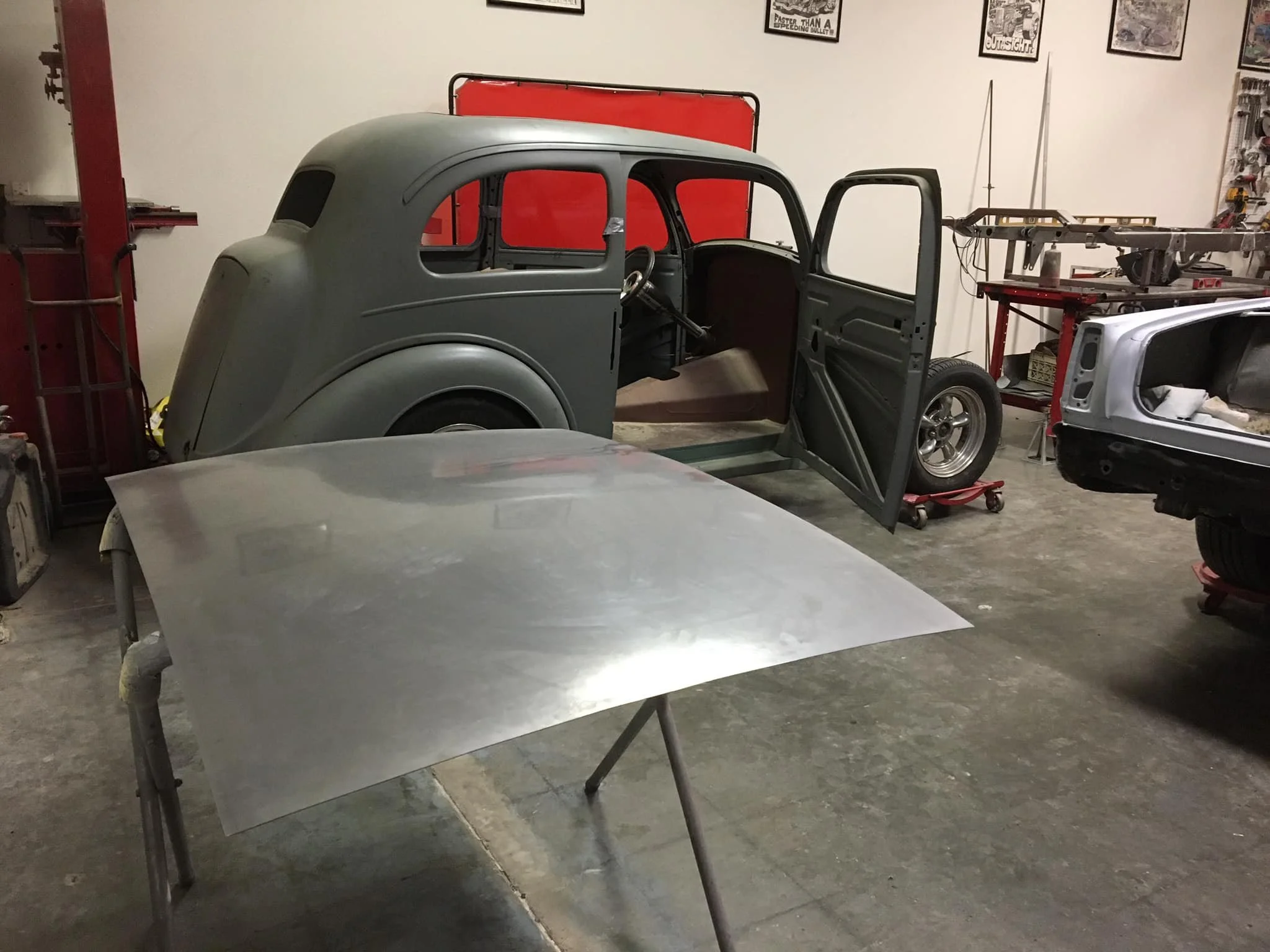 A vintage car body with a gray primer coat and an open door inside a workshop, surrounded by workshop tools and parts, including a metal hood on a stand in the foreground.