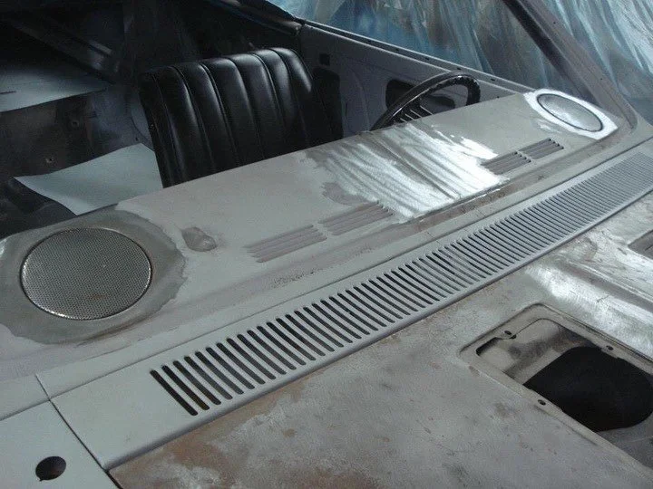 The image shows the interior dashboard and windshield area of an older, possibly vintage, car with a white or light-colored surface, featuring two large circular speakers and a sun visor. The dashboard appears to be dusty or in need of cleaning.