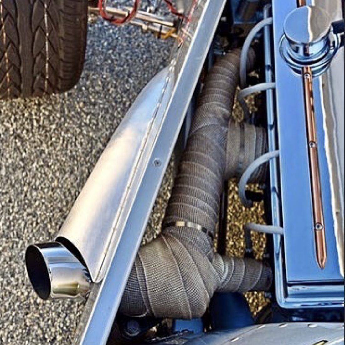 On a gravel surface, a metal table holds a car exhaust pipe and a large flexible hose connected to a metallic box with blue straps.