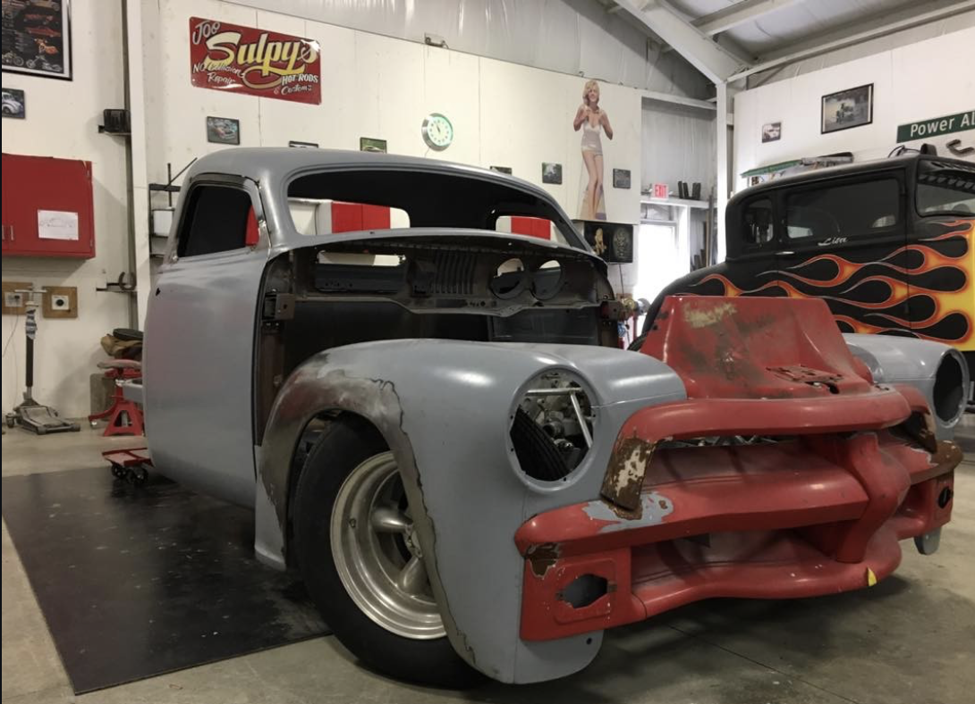 A vintage car body in a garage, partially painted silver with a red front bumper, surrounded by automotive tools and retro signs.