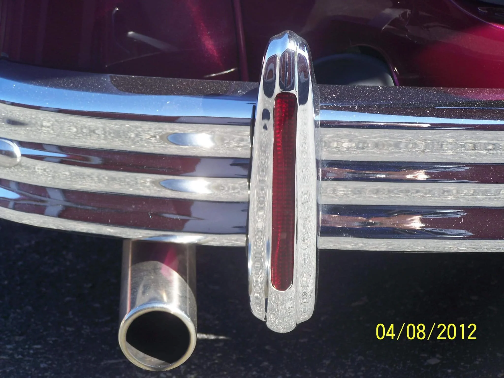 Close-up of the rear end of a classic car with red paint, chrome details, a vertical tail light, and a chrome exhaust pipe, with a date stamp of April 8, 2012.