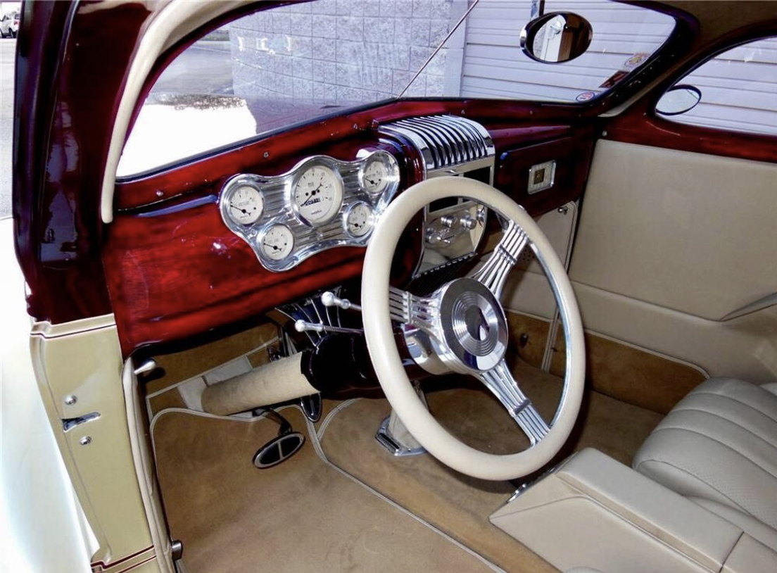 Interior of a vintage car showcasing a wood dashboard with multiple analog gauges, a large steering wheel with chrome accents, and beige leather seats.