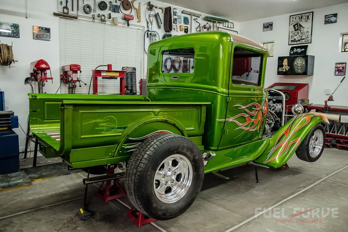 A green classic pickup truck with flame decals being restored in a garage filled with tools and car parts.