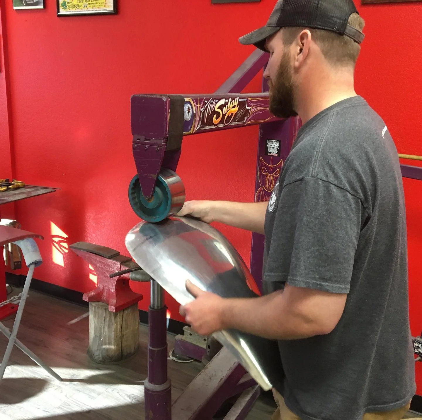 A man wearing a cap and a gray T-shirt polishing a metal skateboard deck with a power tool in a workshop with a red wall.