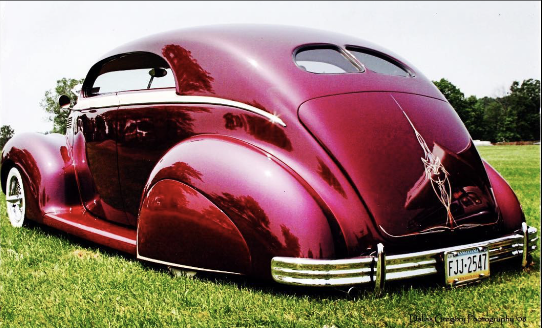 A vintage, custom maroon and black classic car with pinstriping, parked on a grassy field with trees in the background.