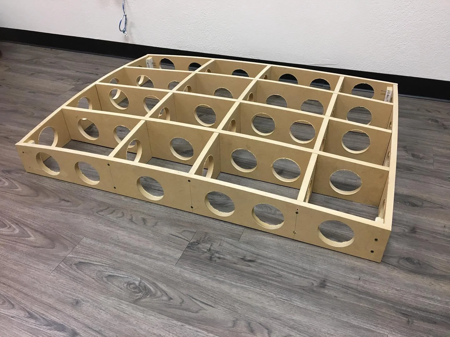 A rectangular wooden frame with multiple circular cutouts, likely a part of furniture or a construction project, placed on a gray wooden floor against a plain white wall.